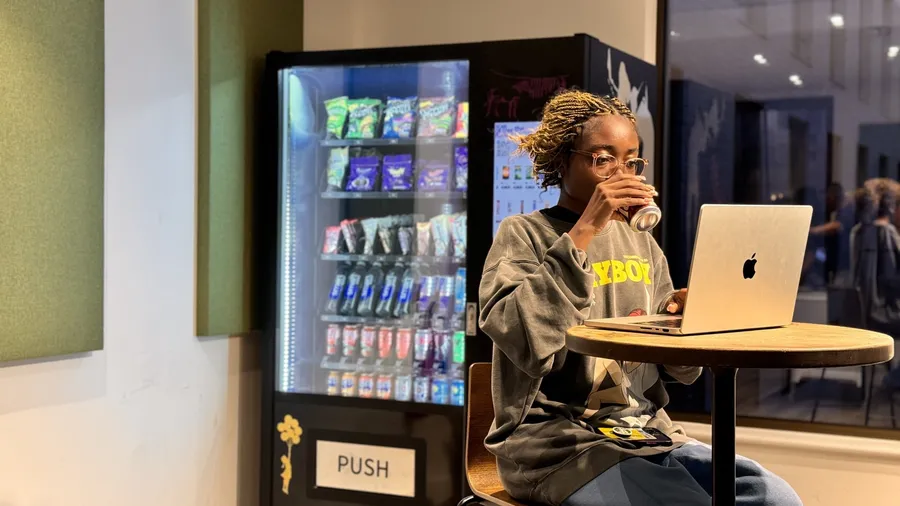 Person working on a laptop beside a Juicy Vend snack and drink machine at Rhodaus Court
