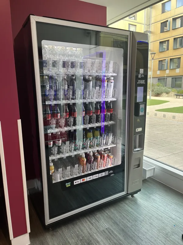 Glass-front drinks machine stocked with water, soft drinks and juices beside a window